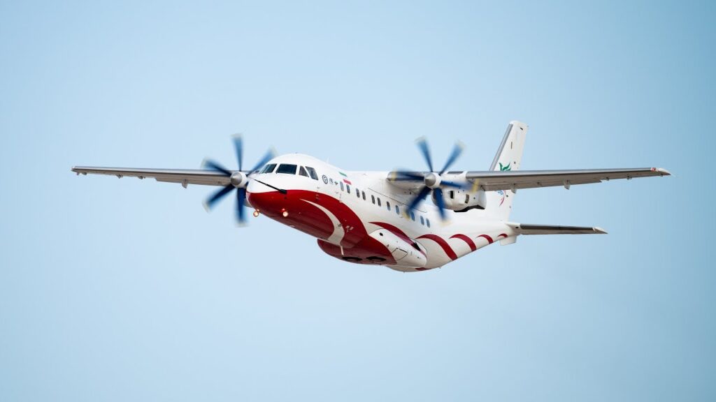 Iran's Simorgh light transport aircraft in flight, a white and livery turboprop plane developed from the IrAn-140, showcasing its high-wing and twin-engine design.